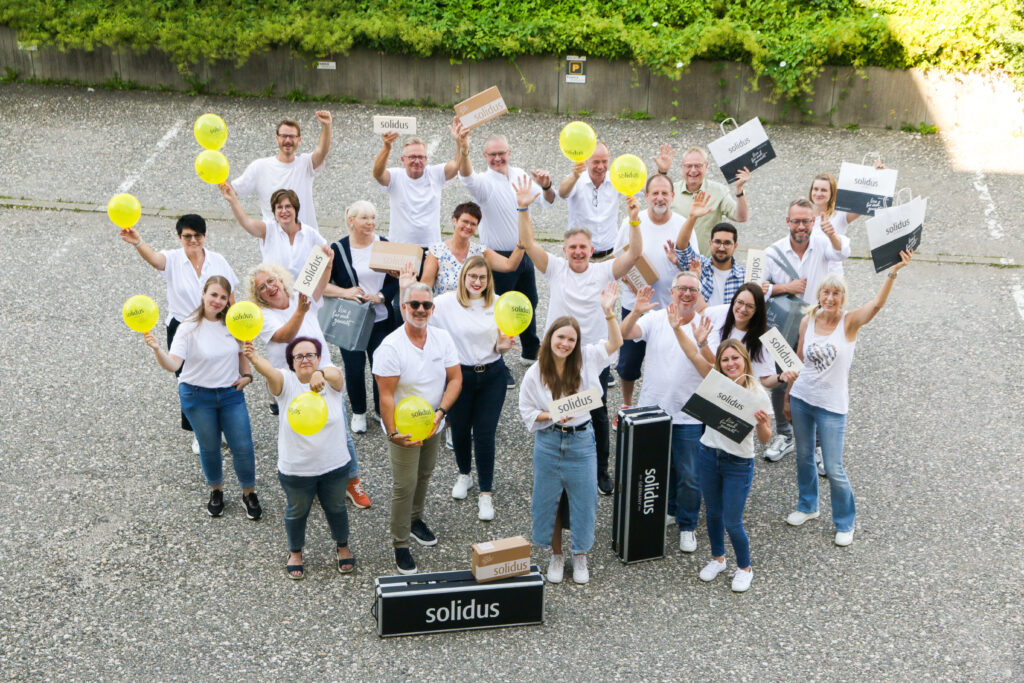 Solidus Teamfoto im Freien mit gelben Ballons, Kartons und Taschen – alle winken in die Kamera