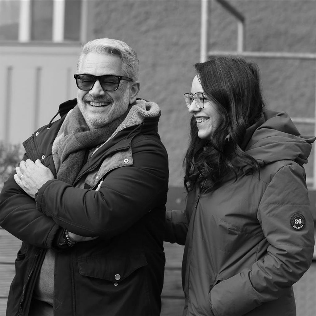 Zwei Mitarbeitende lachen im Freien in Winterjacken, Schwarzweiß