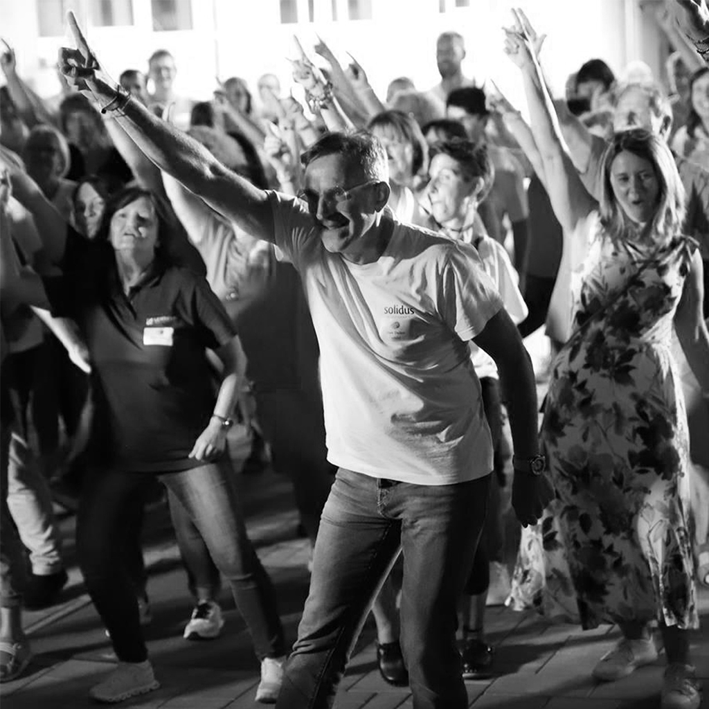 Große Gruppe tanzt synchron, Vordergrund zeigt Mitarbeiter mit erhobenem Arm, Schwarzweiß