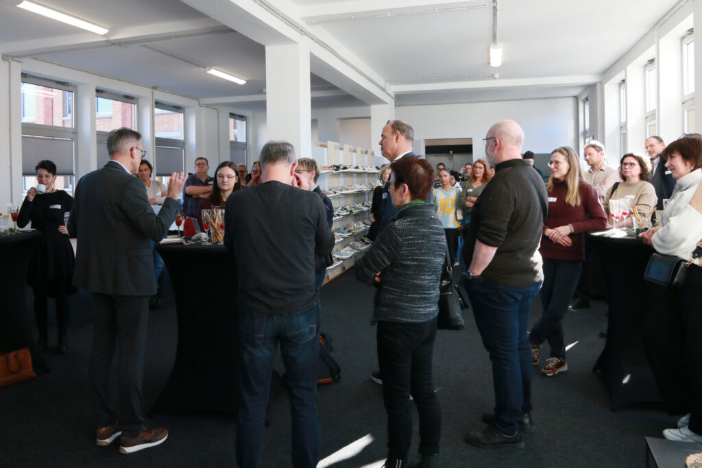 Teilnehmende stehen beim Solidus Come Together im Showroom und hören einer Ansprache zu, im Hintergrund sind Schuhmodelle ausgestellt