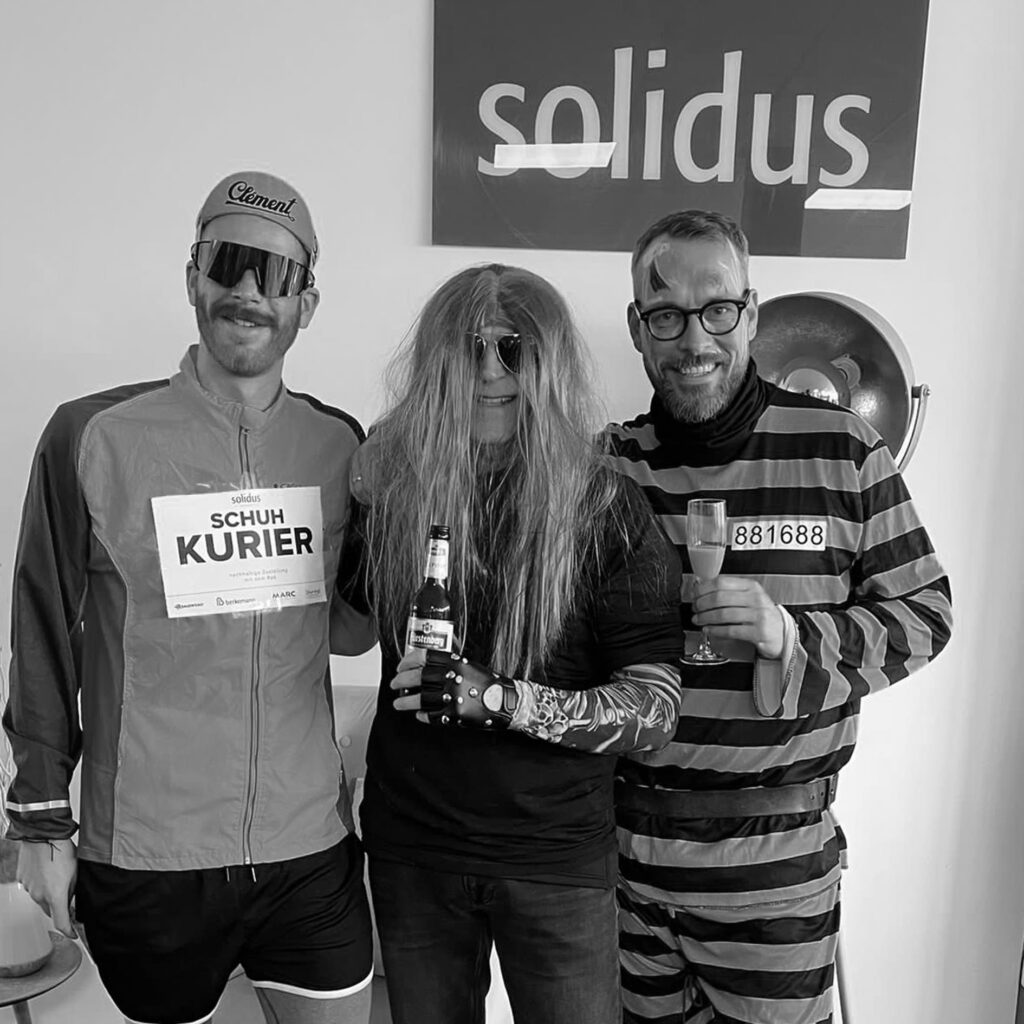Michael Großmann mit zwei Kolleg:innen verkleidet bei der Fasnet im Büro, Solidus Schild im Hintergrund, Kostüm „Schuh Kurier“