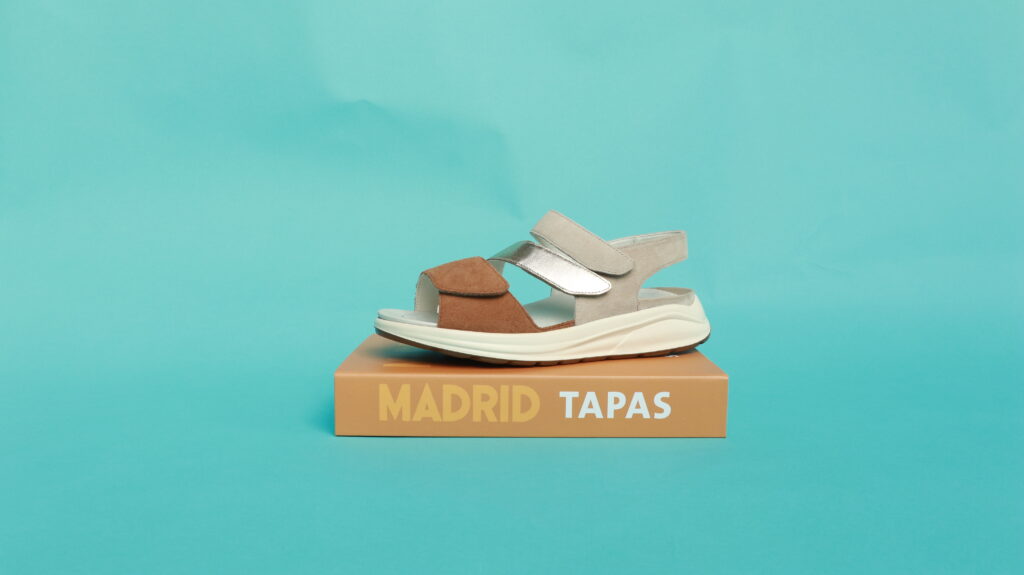 Solidus Komfort-Sandale in Beige mit braunem und silbernem Riemen auf Box „Madrid Tapas“ vor türkisem Hintergrund