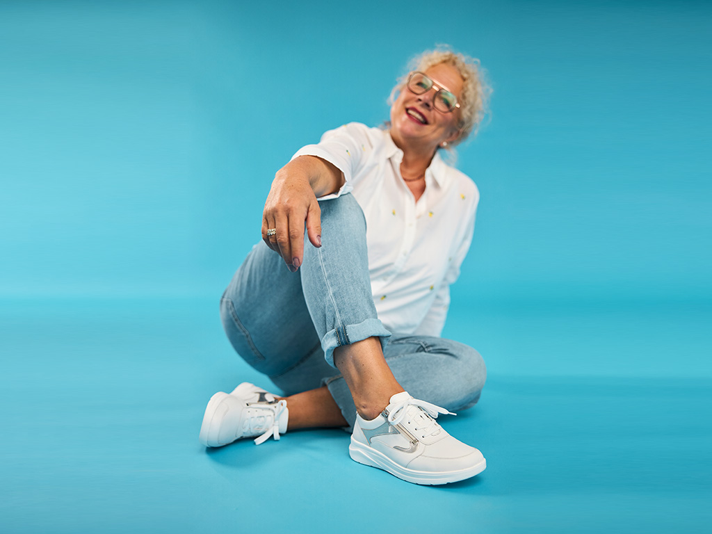 Lächelnde Frau im Studio trägt weiße Solidus Damen-Sneaker und sitzt entspannt vor türkisfarbenem Hintergrund