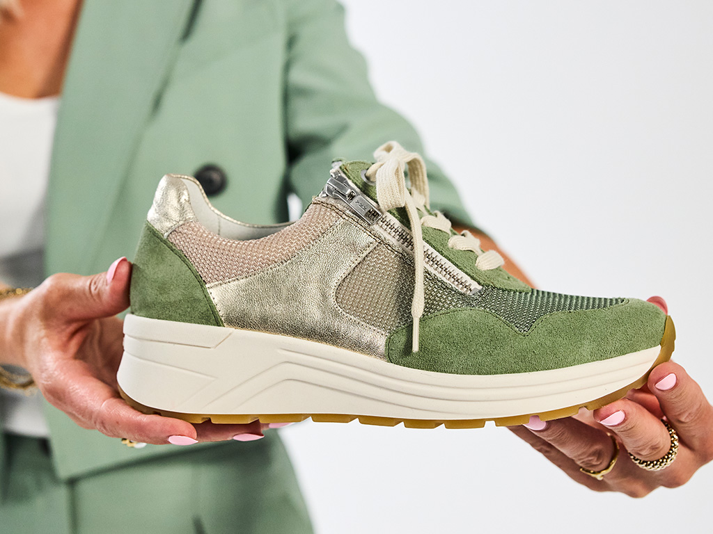 Hände halten grün-goldenen Solidus Damen-Sneaker mit Reißverschluss, Schnürung und profilierter Sohle vor hellem Hintergrund