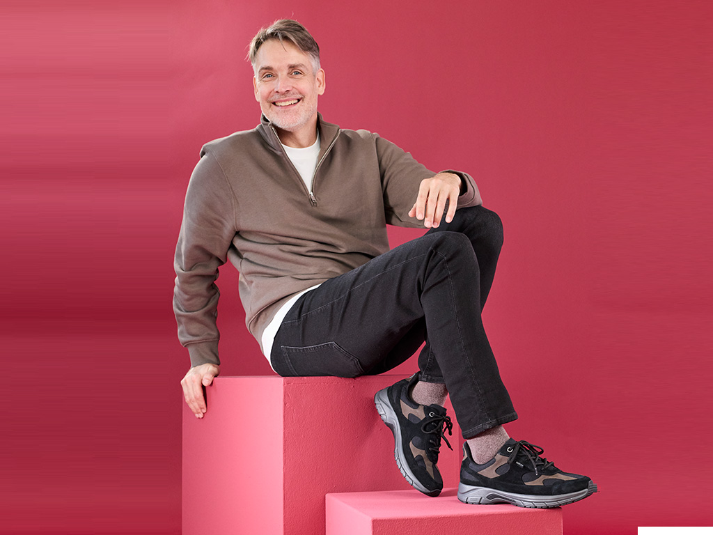 Mann sitzt im Studio vor rotem Hintergrund und trägt sportliche Solidus Herren-Sneaker in Schwarz mit Komfortsohle