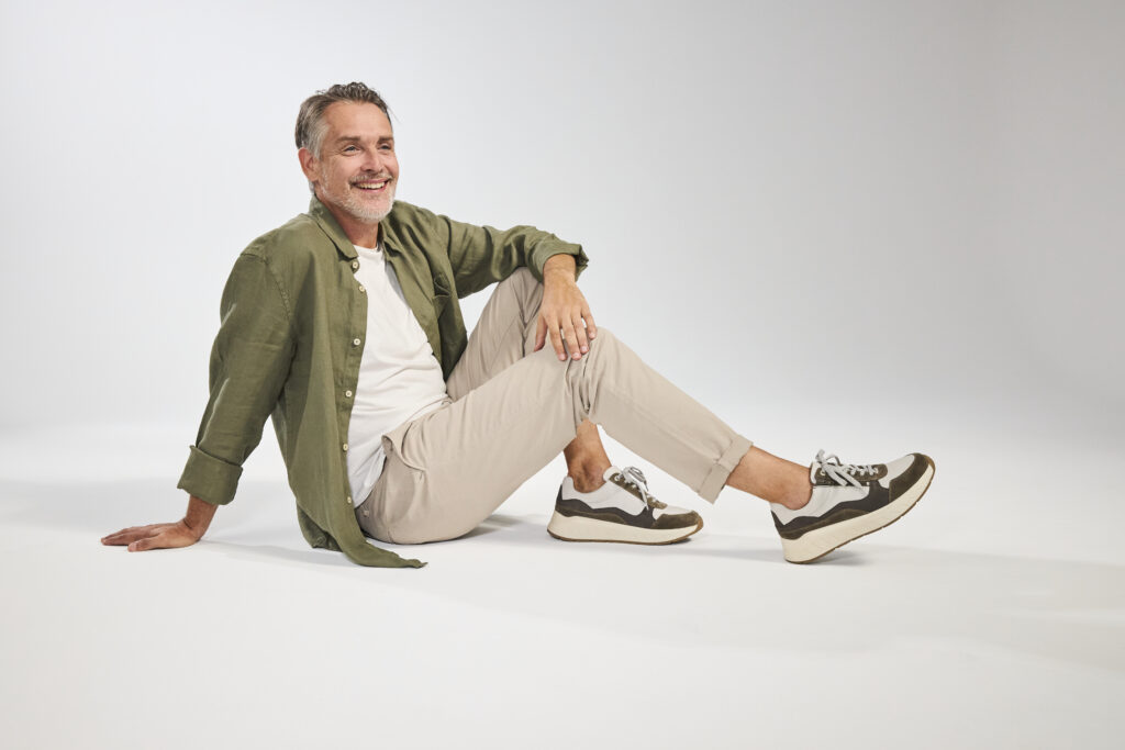 Lächelnder Mann sitzt im Studio und trägt sportliche Solidus Herren-Sneaker in Oliv und Grau